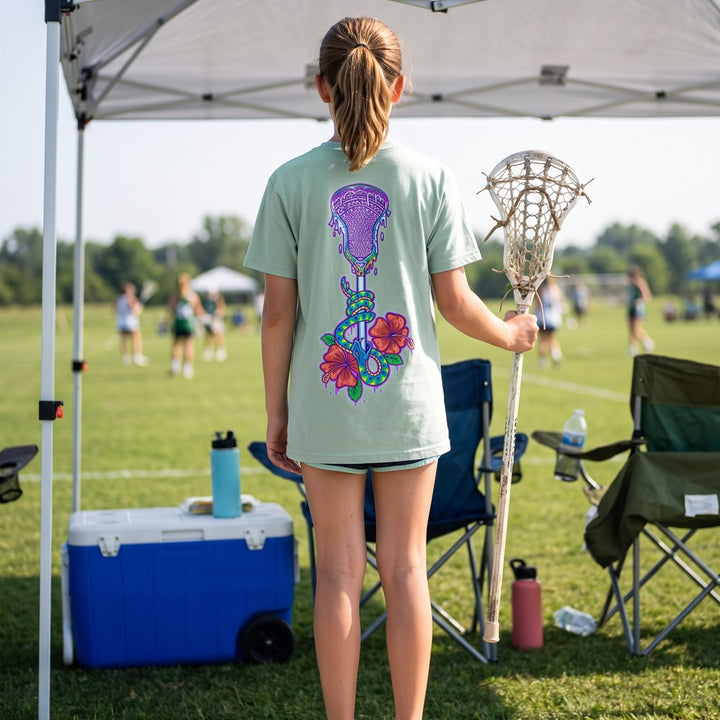 Youth Lax Snake Tee in seafoam worn by a girl at a lacrosse tournament, back view showing the lacrosse stick, snake and hibiscus print