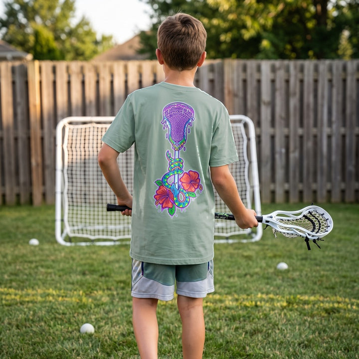 Youth Lax Snake Tee in seafoam worn by a boy in a backyard, back view showing the lacrosse stick, snake and hibiscus print